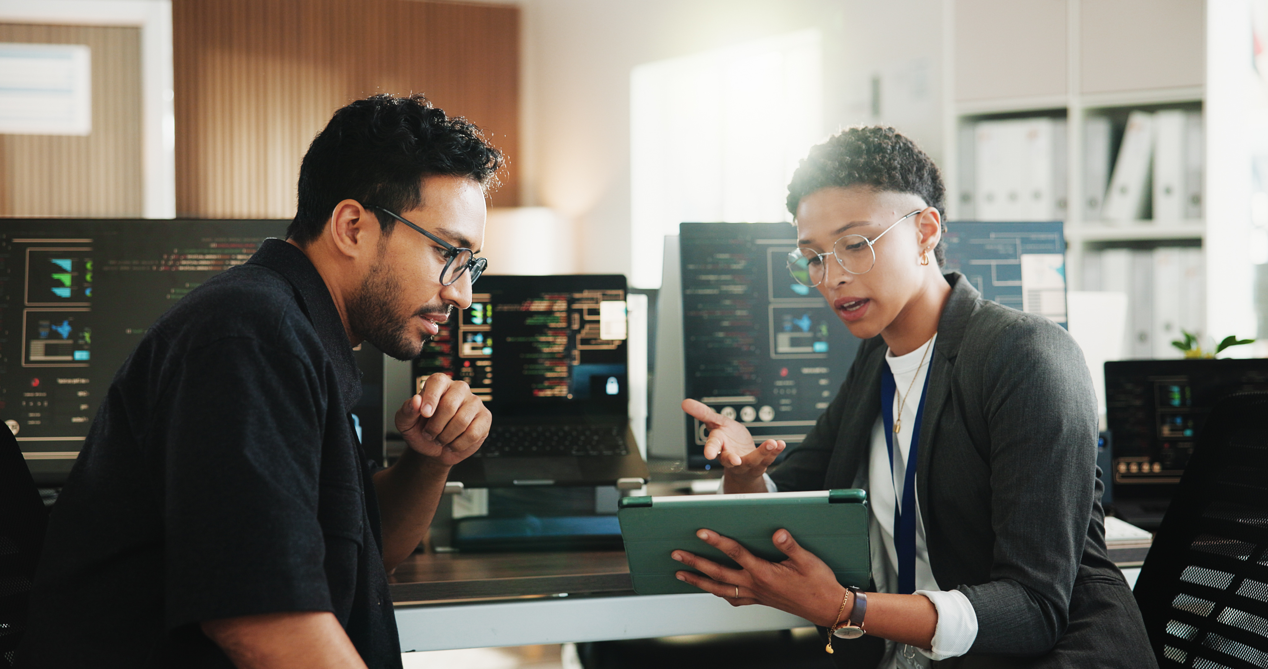 Two professionals reviewing technology on a tablet in a modern office with code on monitors behind them